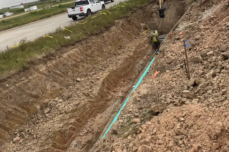 A deep trench with green pipe bedded in it, running beside a live roadway